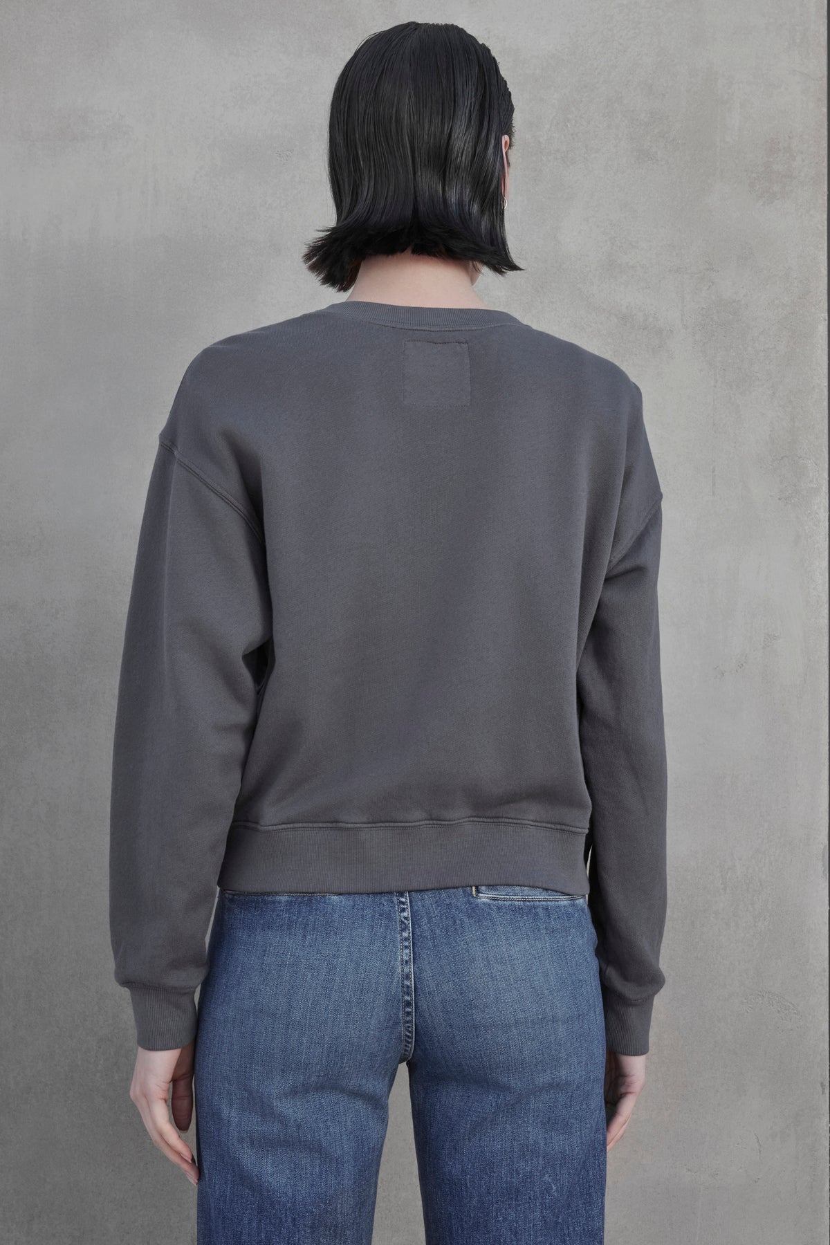   A person with short dark hair is standing with their back turned, wearing a dark gray YNEZ SWEATSHIRT made from organic fleece by Velvet by Jenny Graham and blue jeans against a plain backdrop. 