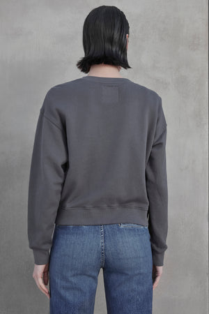 A person with short dark hair is standing with their back turned, wearing a dark gray YNEZ SWEATSHIRT made from organic fleece by Velvet by Jenny Graham and blue jeans against a plain backdrop.