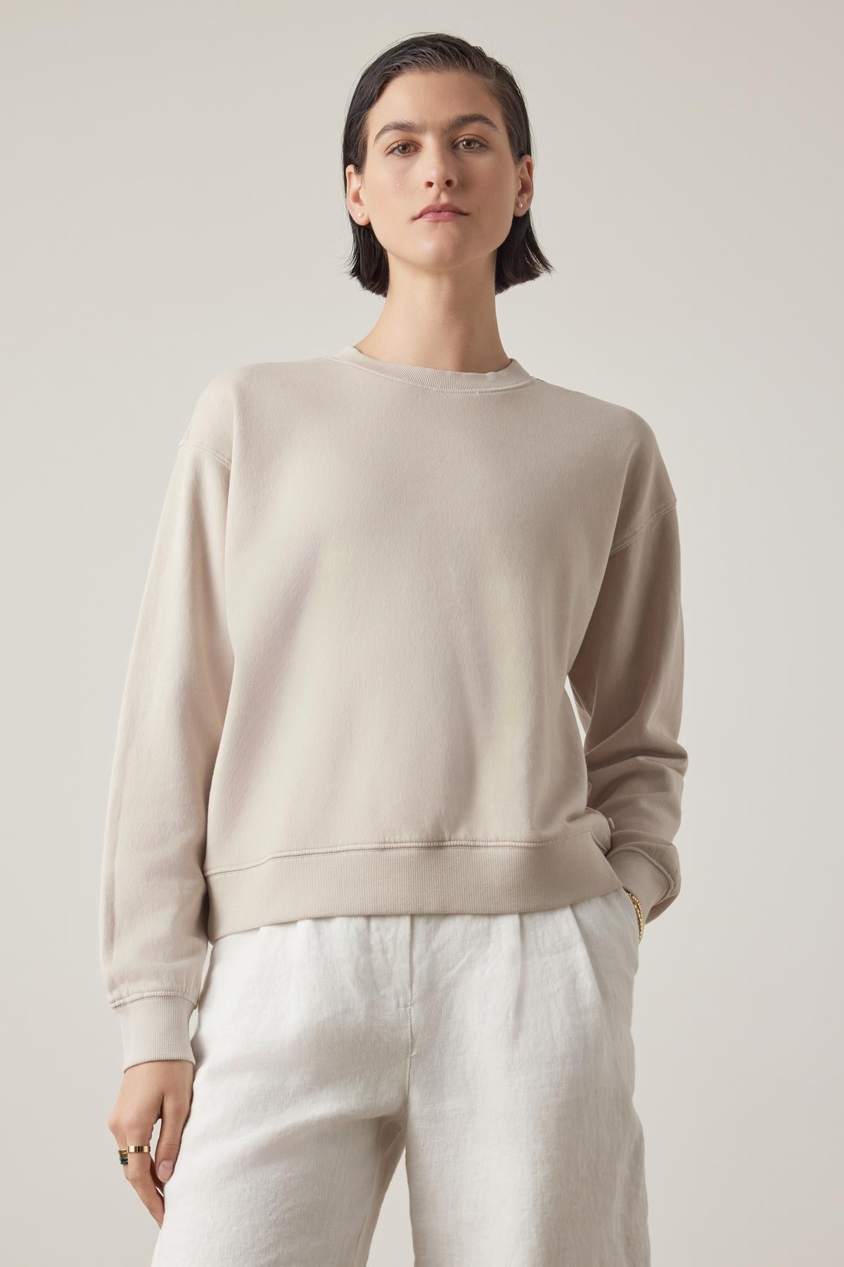   Woman in a Velvet by Jenny Graham Ynez Sweatshirt and white pants standing against a plain background, looking directly at the camera. 