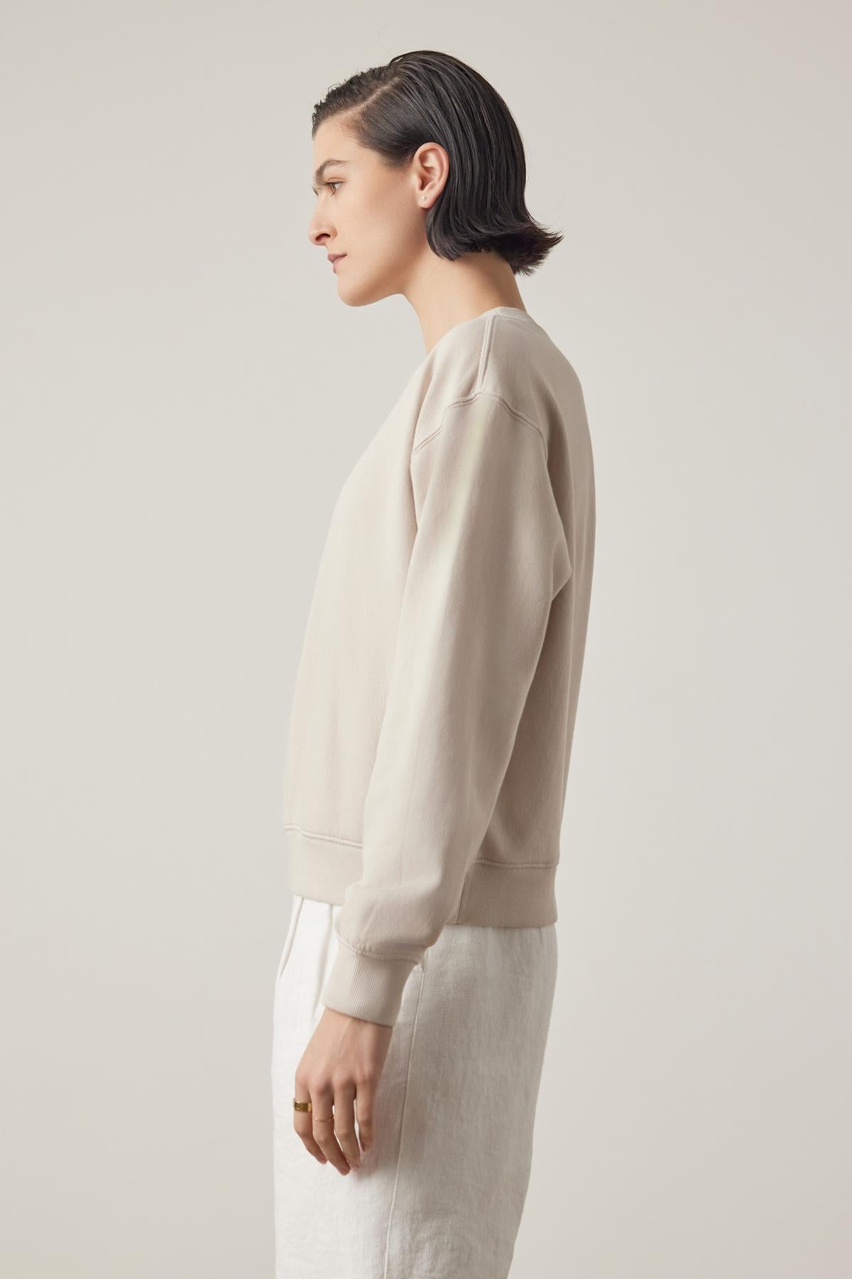   Profile view of a person with slicked-back hair wearing a beige YNEZ SWEATSHIRT by Velvet by Jenny Graham and white pants against a light background. 
