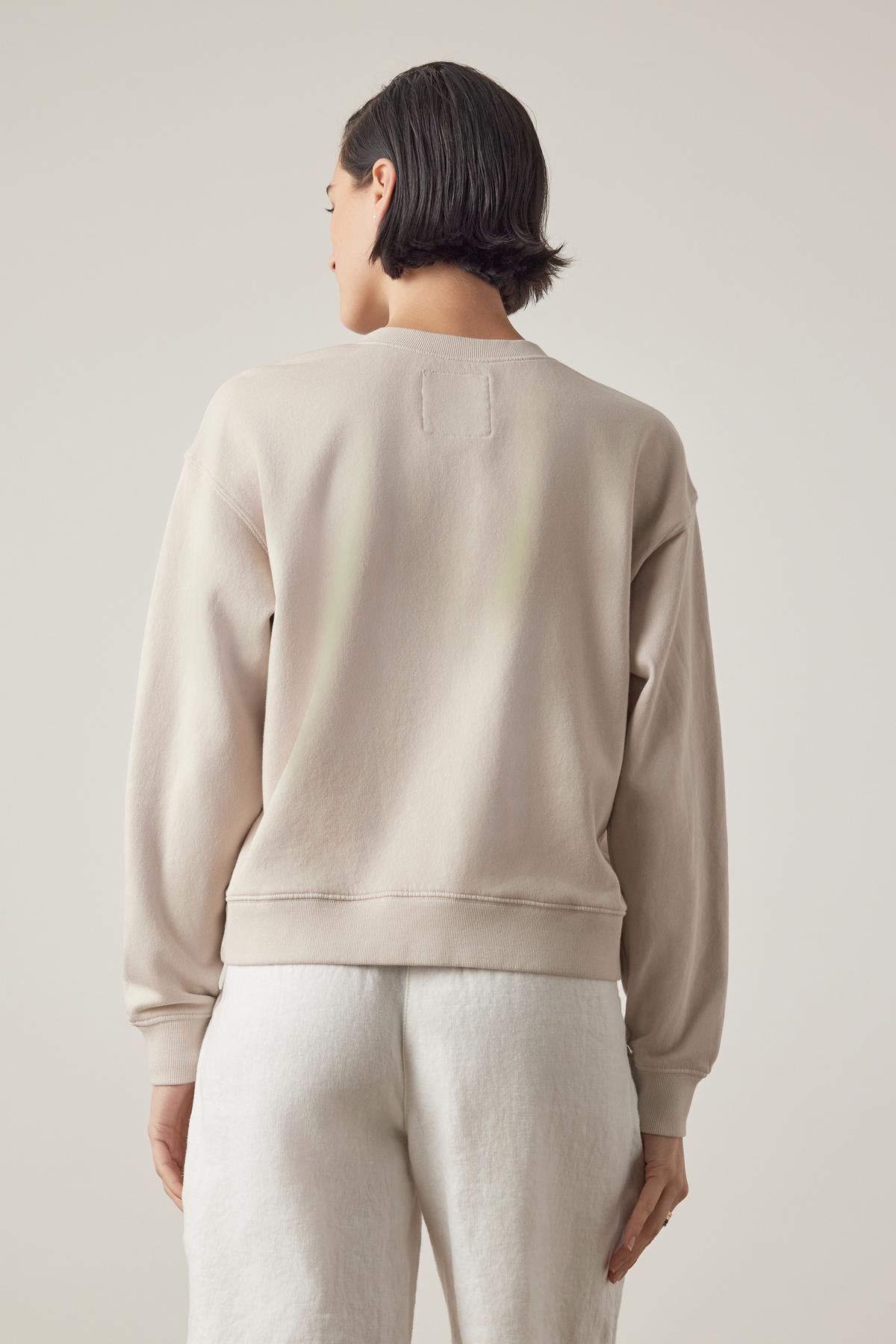 Woman from behind wearing a beige organic YNEZ Sweatshirt by Velvet by Jenny Graham and white pants against a neutral background.-36863307710657