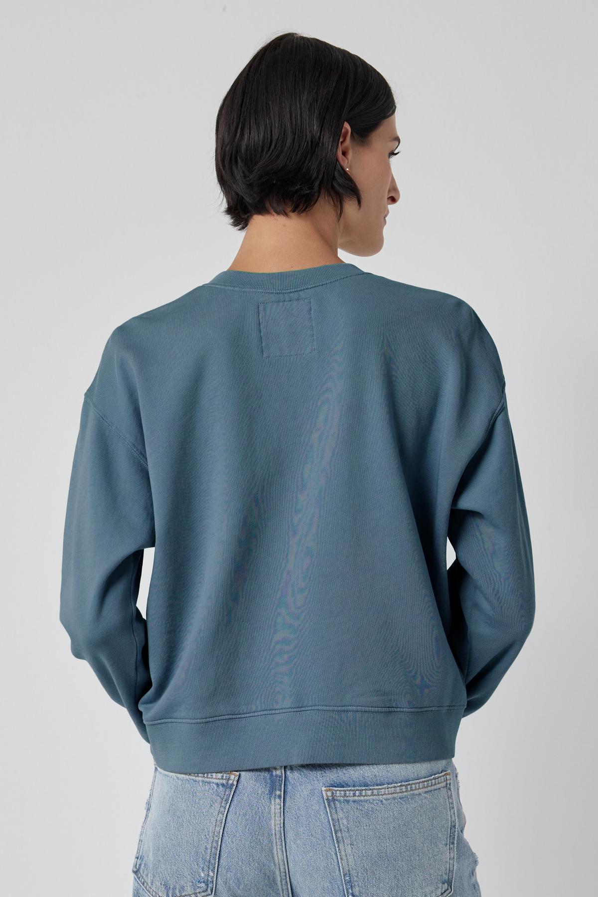 The back view of a woman wearing a blue Ynez Sweatshirt by Velvet by Jenny Graham with a dropped shoulder.-35721183756481