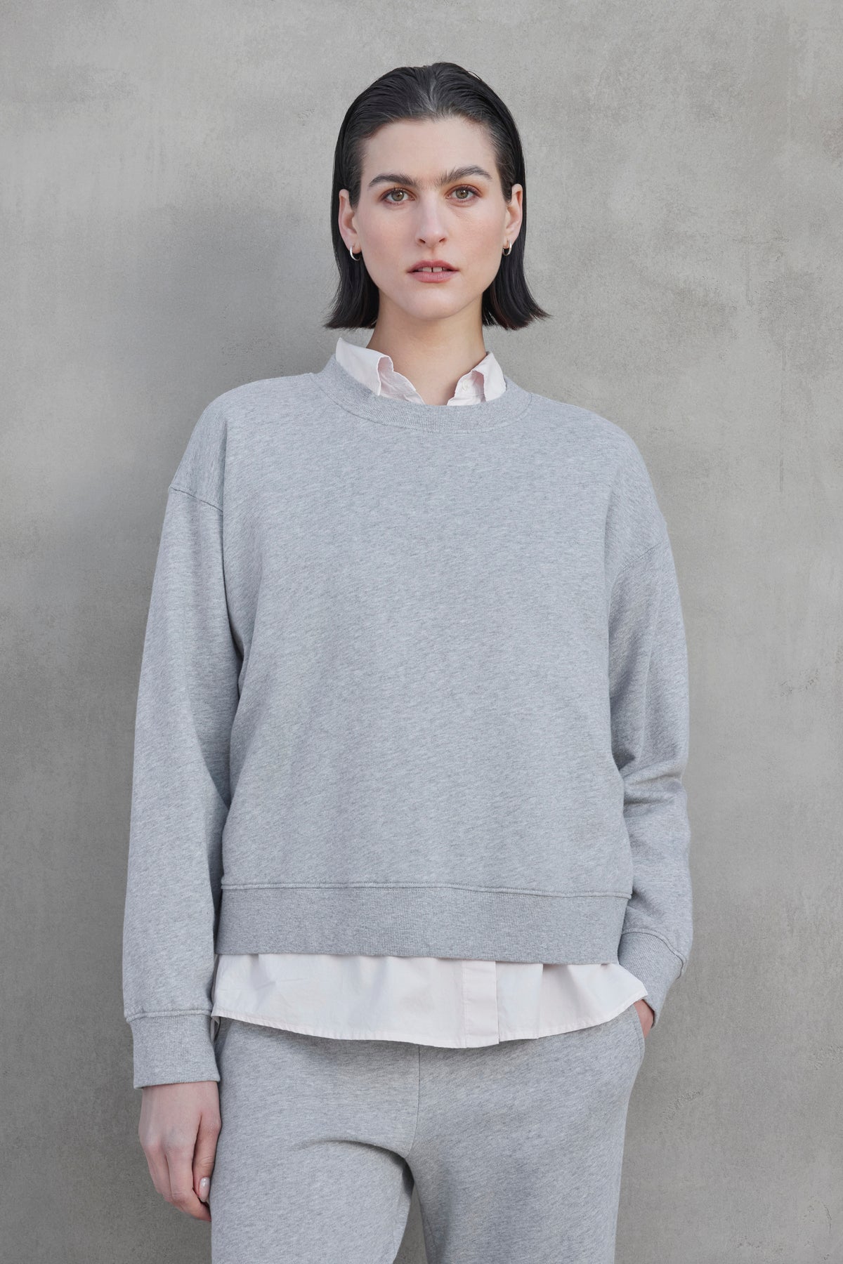   A person with short dark hair wears the YNEZ SWEATSHIRT by Velvet by Jenny Graham, made from organic cotton with exaggerated seams, over a white shirt and gray pants, standing against a plain gray background. 