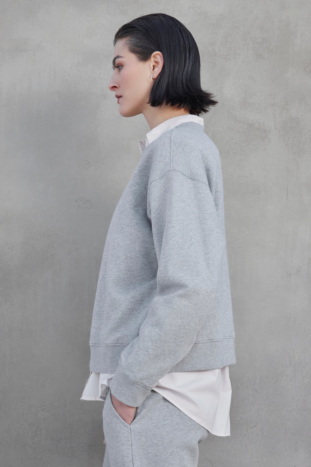   A person with short black hair wearing a YNEZ SWEATSHIRT by Velvet by Jenny Graham, made from organic cotton, and a white shirt stands in profile against a gray concrete wall. 