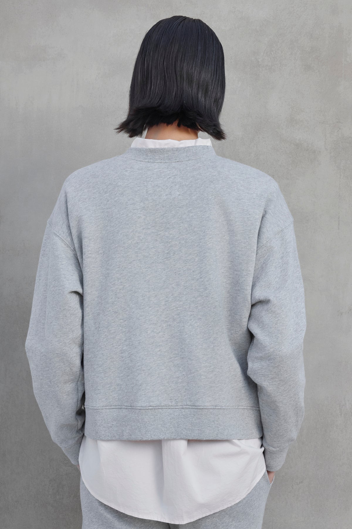   The individual with short black hair is facing a textured gray wall, dressed in the YNEZ SWEATSHIRT by Velvet by Jenny Graham, layered over a white shirt. 