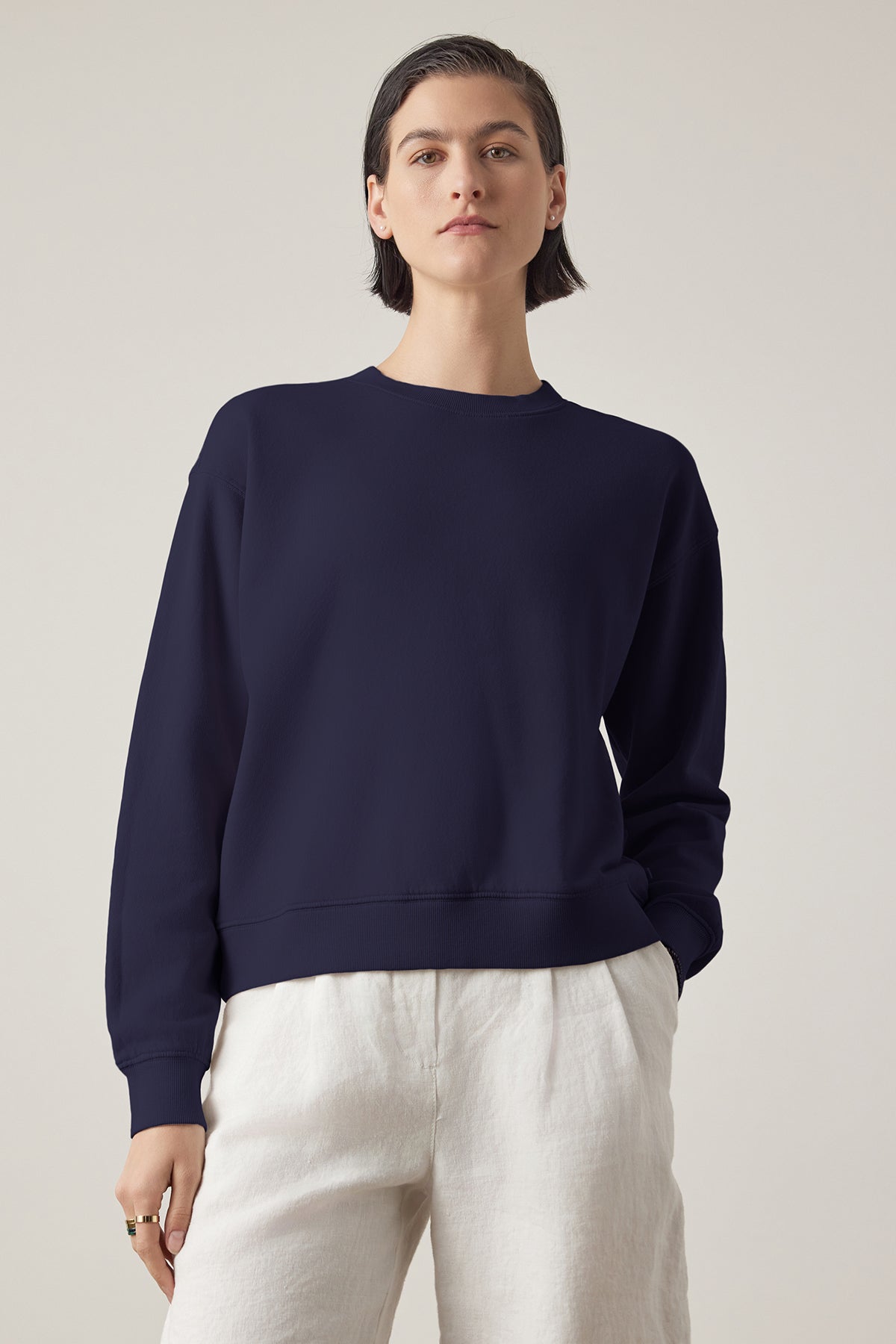   Woman in a Velvet by Jenny Graham Ynez Sweatshirt and white pants standing against a neutral background. 