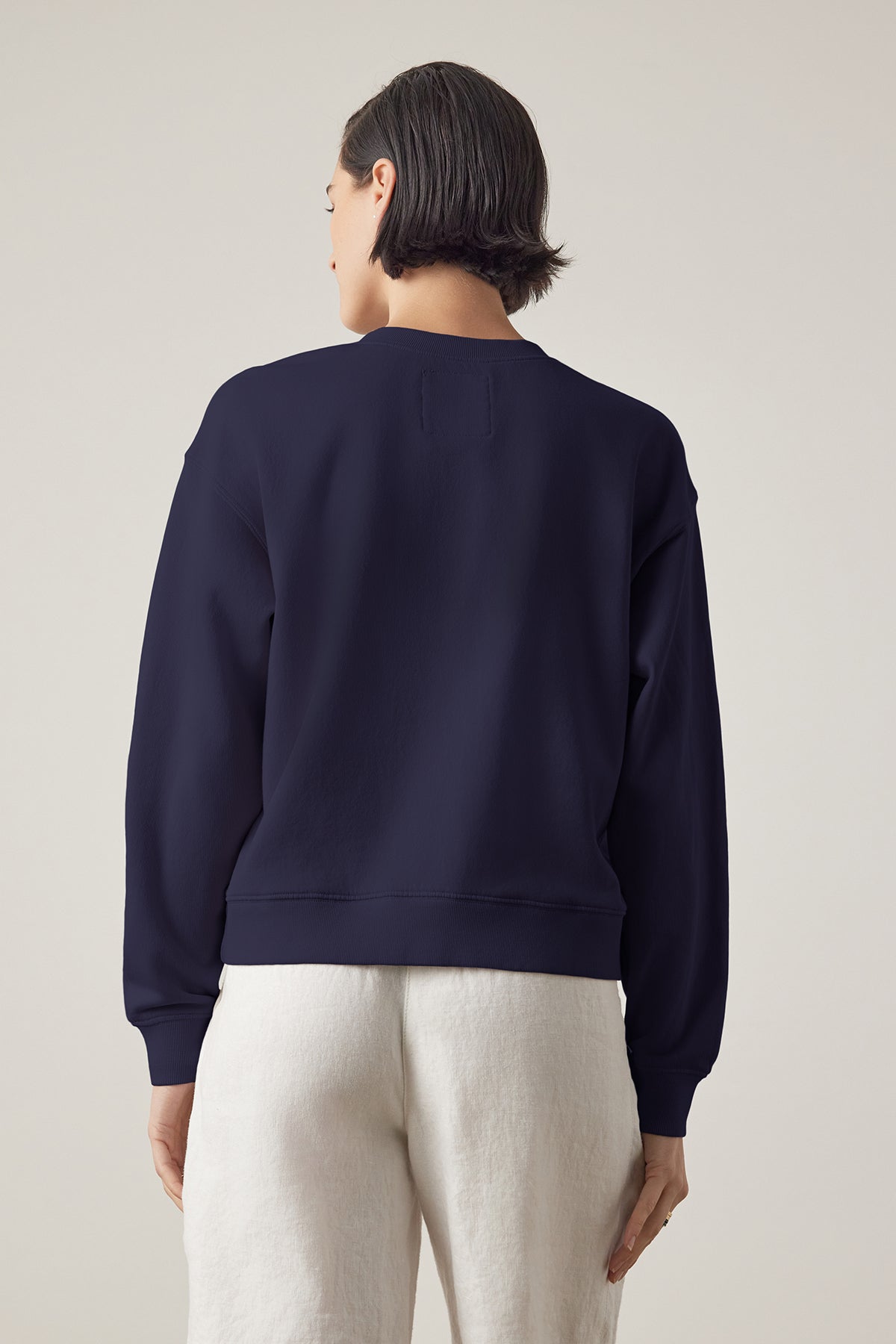   A woman seen from behind wearing a YNEZ SWEATSHIRT made by Velvet by Jenny Graham, made of organic fleece and white pants against a neutral background. 