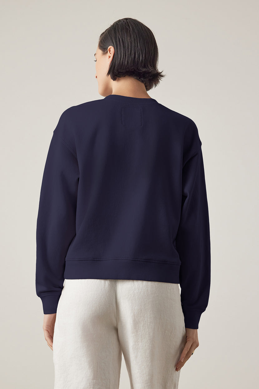 A woman seen from behind wearing a YNEZ SWEATSHIRT made by Velvet by Jenny Graham, made of organic fleece and white pants against a neutral background.