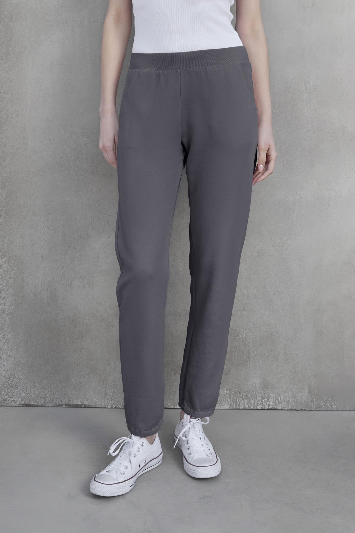 A person wearing Velvet by Jenny Graham's ZUMA SWEATPANT, a white tank top, and white sneakers stands against a gray concrete wall.-37817336496321