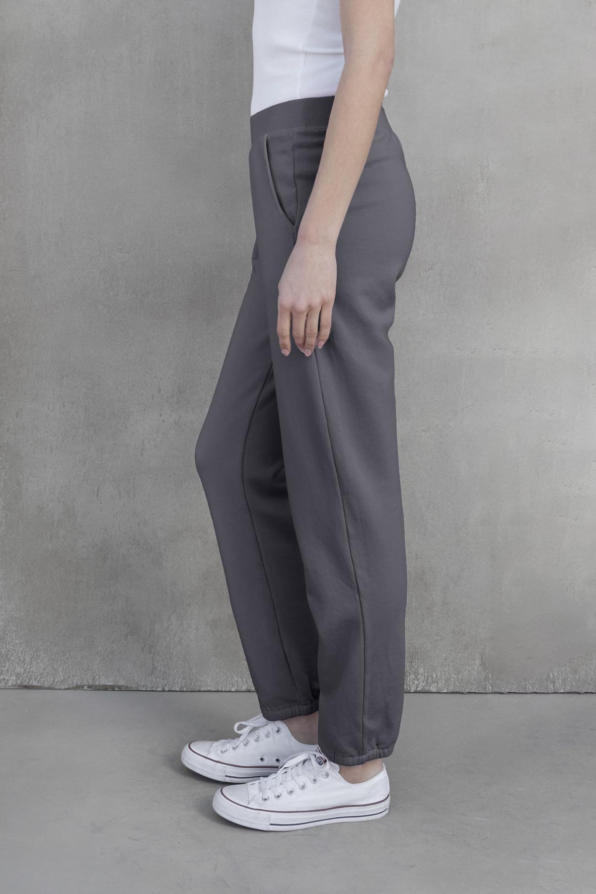   A person standing sideways, wearing a white top, the ZUMA SWEATPANT in gray by Velvet by Jenny Graham, and white sneakers, against a concrete wall background. 