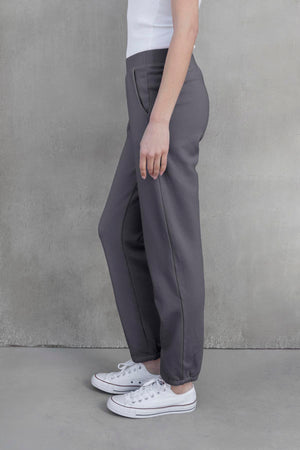 A person standing sideways, wearing a white top, the ZUMA SWEATPANT in gray by Velvet by Jenny Graham, and white sneakers, against a concrete wall background.