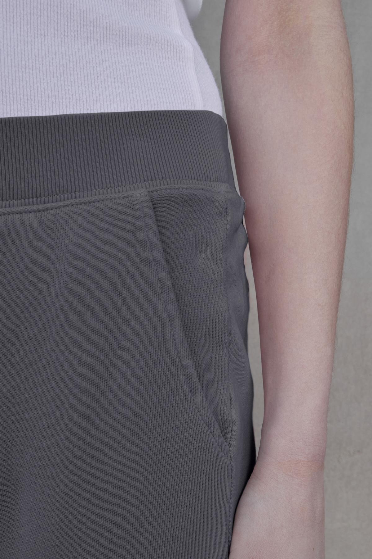   The image shows a close-up of a person wearing a white shirt and the ZUMA SWEATPANT by Velvet by Jenny Graham, made from dark gray organic cotton, with their hand resting in the pants pocket. 