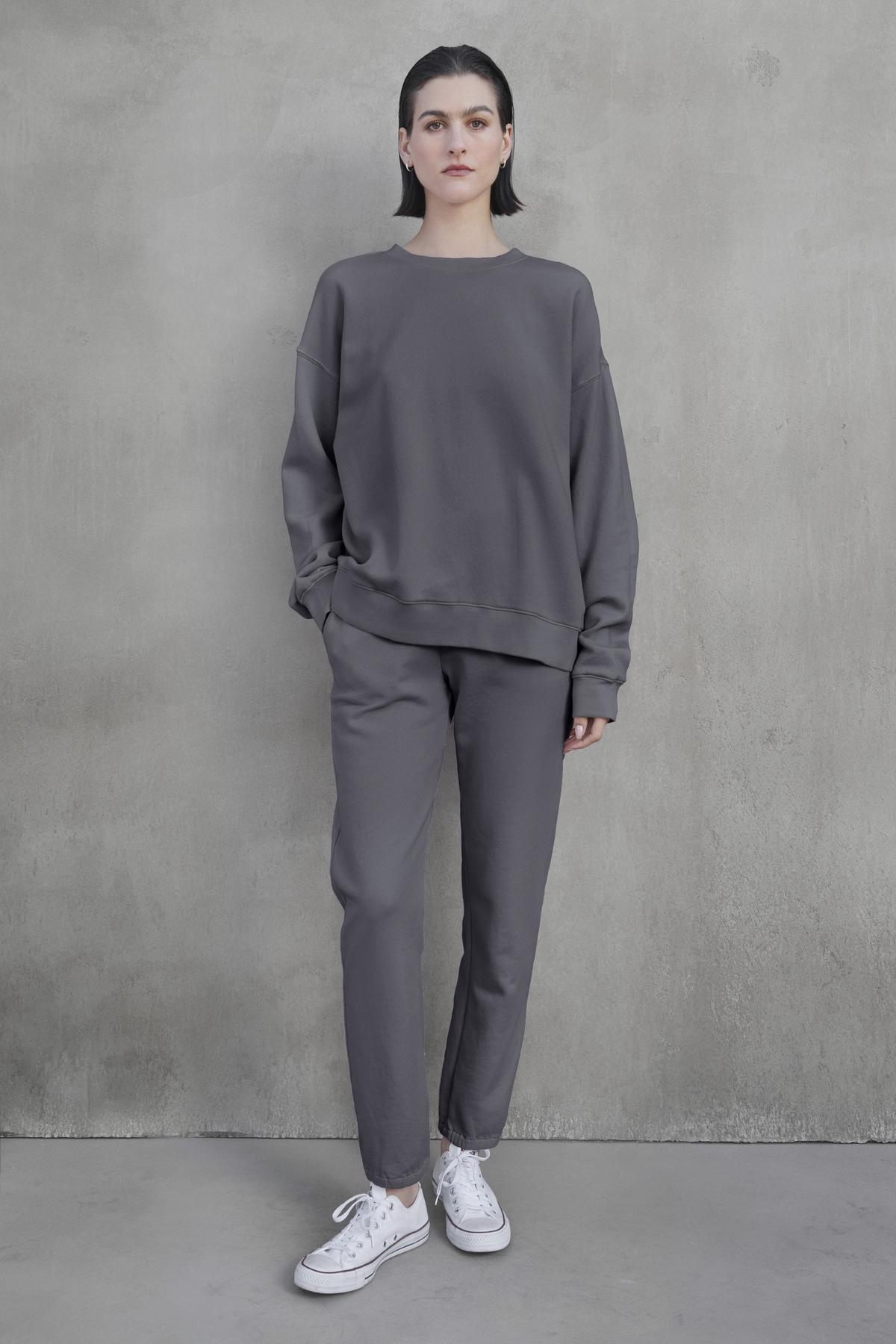   A person stands against a gray concrete wall, wearing the ZUMA SWEATPANT by Velvet by Jenny Graham and a matching loose, gray sweatshirt, paired with white sneakers. They have short, dark hair and a neutral expression, embodying sustainable fashion. 