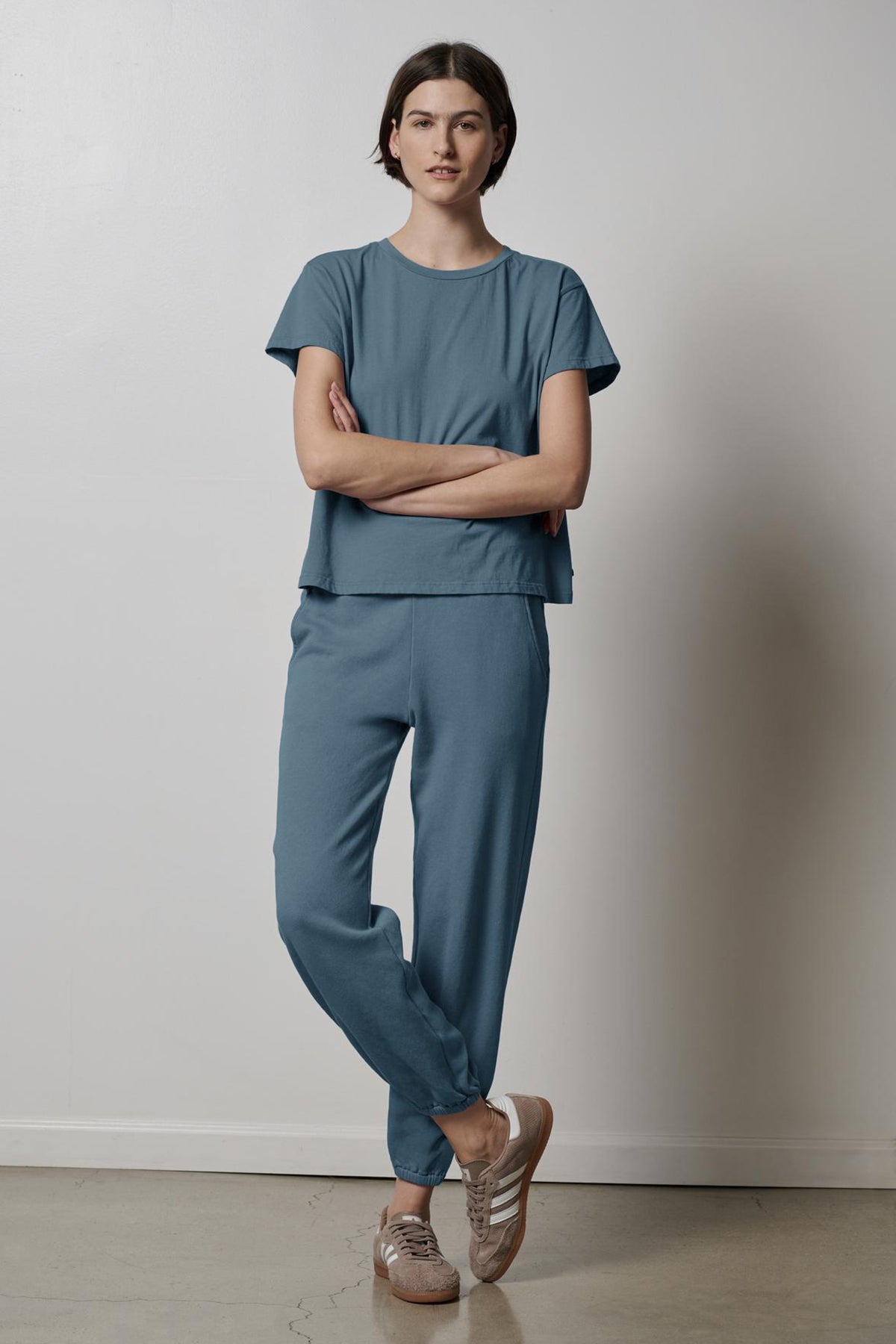 A woman standing with arms crossed wearing a casual blue t-shirt and Velvet by Jenny Graham ZUMA SWEATPANT outfit with brown sneakers.-36594727977153