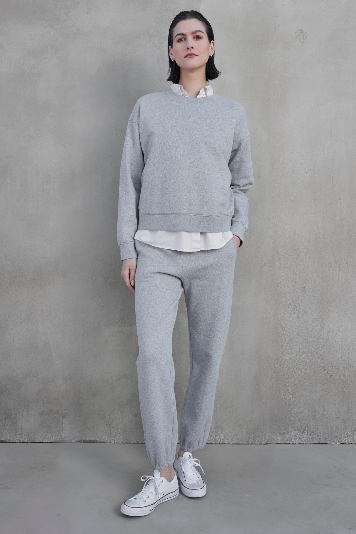   A person stands against a concrete wall, wearing the light gray YNEZ SWEATSHIRT by Velvet by Jenny Graham with exaggerated seams, matching sweatpants, a white shirt, and white sneakers. 