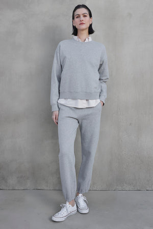 A person stands against a concrete wall, wearing the light gray YNEZ SWEATSHIRT by Velvet by Jenny Graham with exaggerated seams, matching sweatpants, a white shirt, and white sneakers.