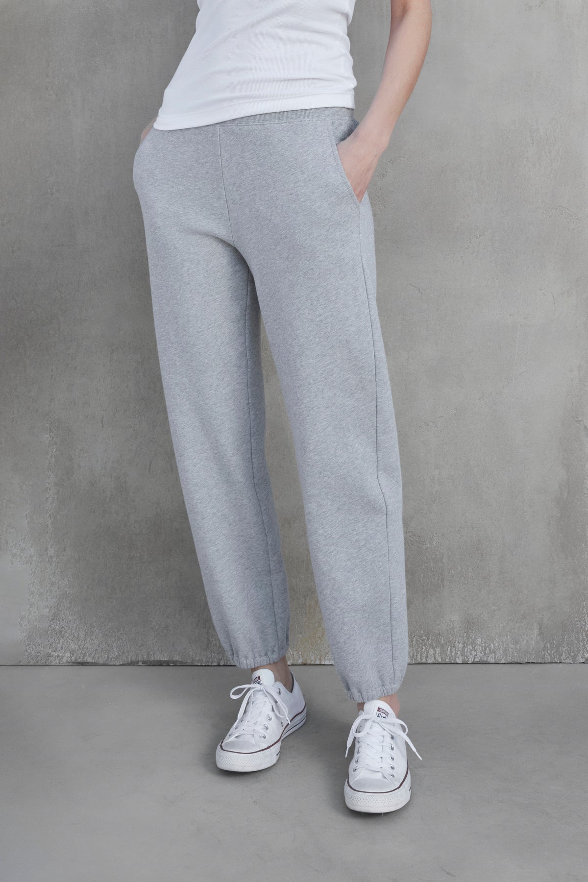   Person wearing a white t-shirt, ZUMA SWEATPANT gray joggers made of organic cotton by Velvet by Jenny Graham with hands in pockets, and white sneakers, standing against a gray concrete wall. 