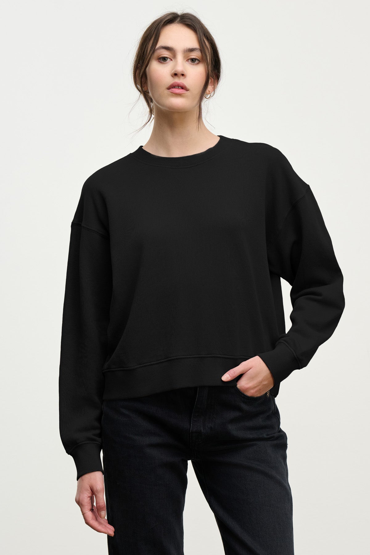 A woman stands against a light background, looking at the camera with one hand in her pocket, wearing Velvet by Graham & Spencer’s YOKO SWEATSHIRT in cotton fleece and black jeans.