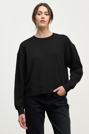 A woman stands against a light background, looking at the camera with one hand in her pocket, wearing Velvet by Graham & Spencer’s YOKO SWEATSHIRT in cotton fleece and black jeans.