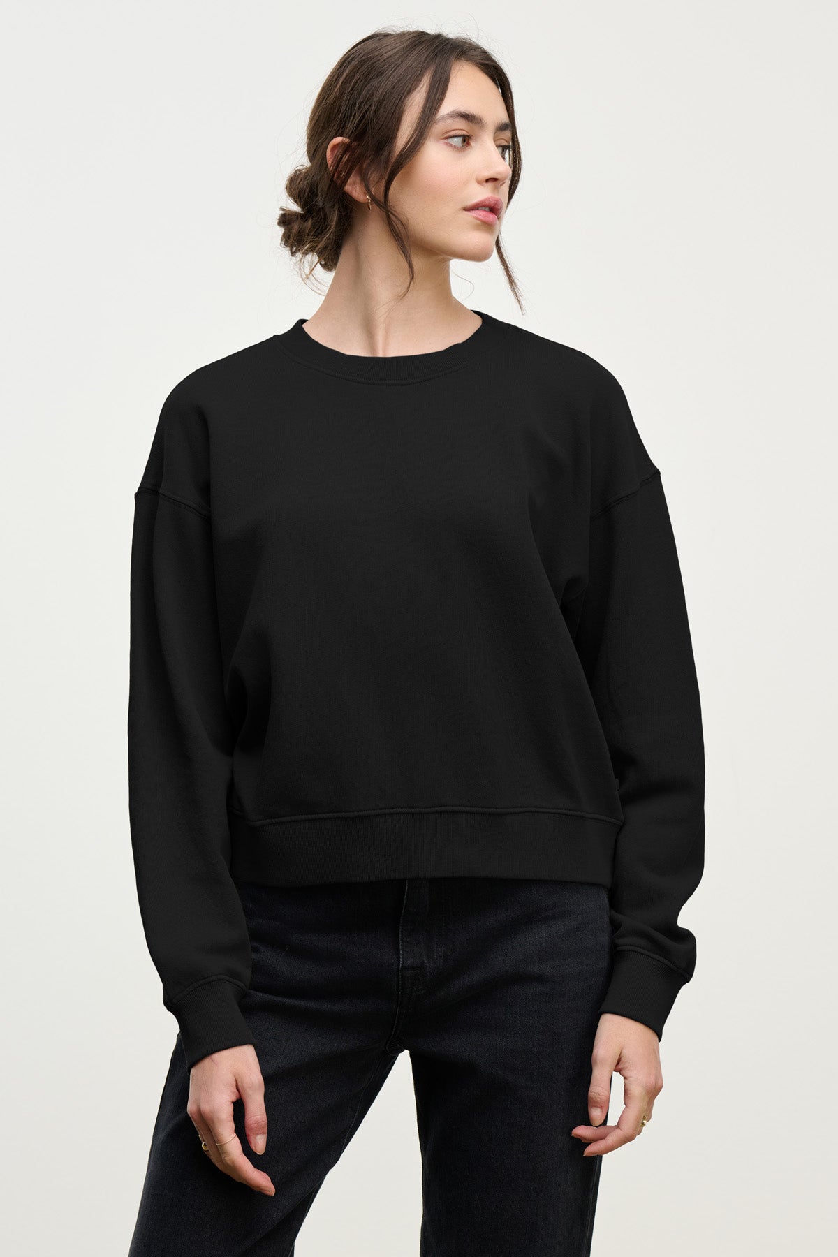 A woman stands against a plain background in the relaxed YOKO SWEATSHIRT by Velvet by Graham & Spencer, made of cotton fleece, paired with black jeans and looking slightly to the side.-40431126675649