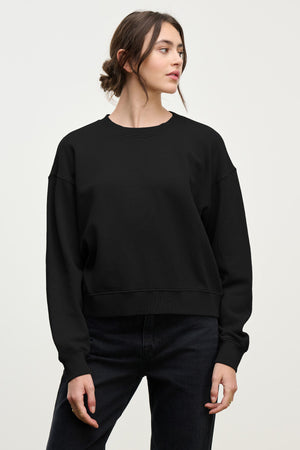A woman stands against a plain background in the relaxed YOKO SWEATSHIRT by Velvet by Graham & Spencer, made of cotton fleece, paired with black jeans and looking slightly to the side.