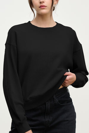 A woman models the YOKO SWEATSHIRT by Velvet by Graham & Spencer in a relaxed fit with black jeans, standing against a light background and holding up one side of the cotton fleece sweatshirt.