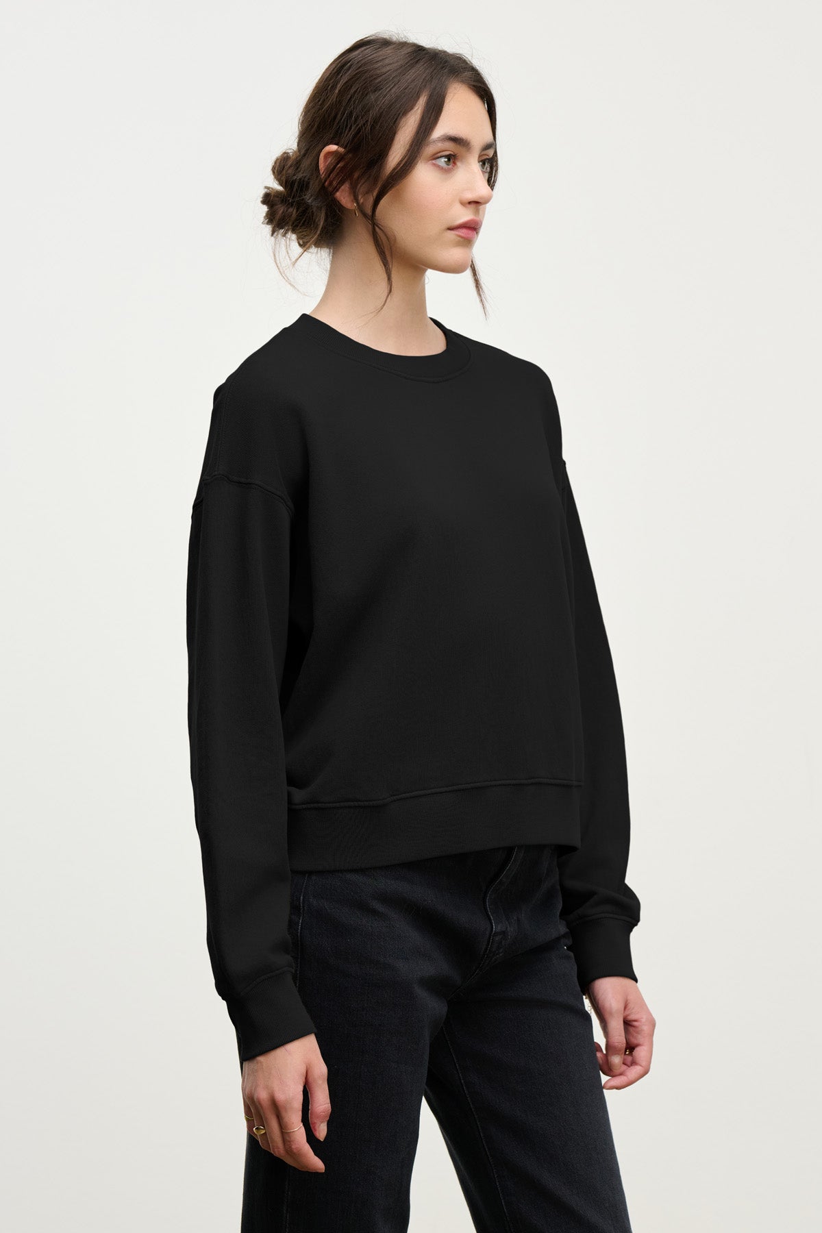 A woman with dark hair in a low bun wears the Velvet by Graham & Spencer YOKO SWEATSHIRT in soft black cotton fleece with black jeans, standing against a plain light background.