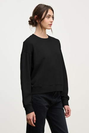 A woman with dark hair in a low bun wears the Velvet by Graham & Spencer YOKO SWEATSHIRT in soft black cotton fleece with black jeans, standing against a plain light background.