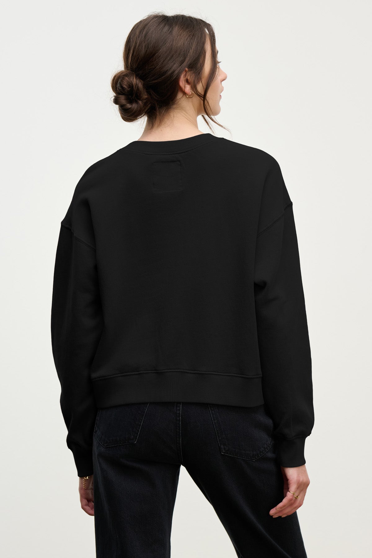 A woman with brown hair in a low bun is seen from the back against a plain light background, wearing the Velvet by Graham & Spencer YOKO SWEATSHIRT in soft cotton fleece with dark jeans for an effortlessly relaxed look.-40431126577345