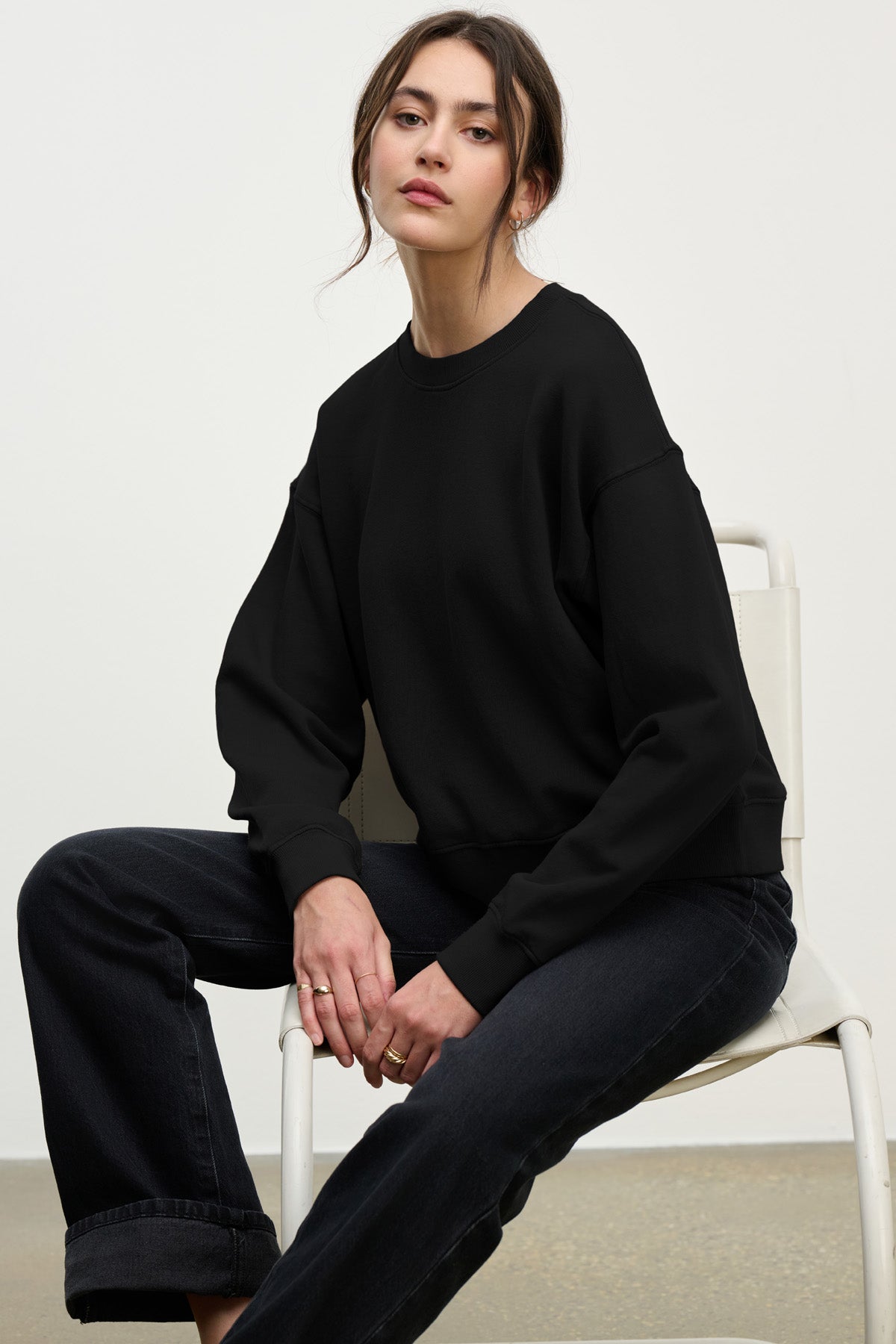A woman sits on a white chair wearing the YOKO SWEATSHIRT in black cotton fleece by Velvet by Graham & Spencer with matching jeans, her hair tied back and a calm expression mirroring the outfit’s relaxed silhouette.