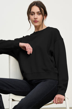 A woman with dark hair in a low bun sits on a chair, wearing the YOKO SWEATSHIRT by Velvet by Graham & Spencer in soft black cotton fleece and dark jeans, looking directly at the camera.