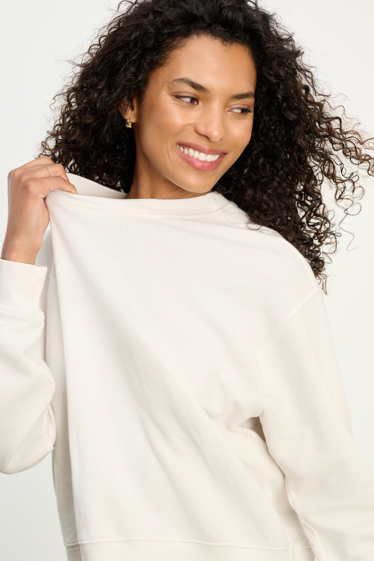 A woman with curly hair smiles and looks to the side, holding the collar of her light YOKO SWEATSHIRT by Velvet by Graham & Spencer, highlighting its relaxed fit.