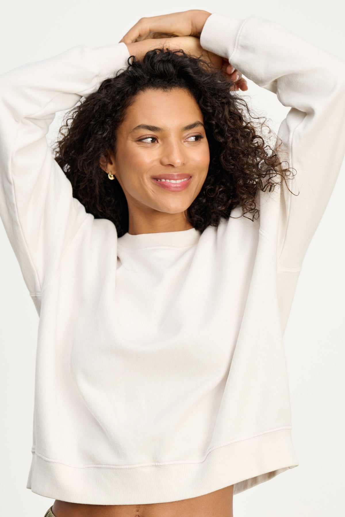 A person with curly hair smiles and looks to the side while wearing a light YOKO SWEATSHIRT in cotton fleece by Velvet by Graham & Spencer, arms raised above their head, showing off its relaxed silhouette against a plain background.