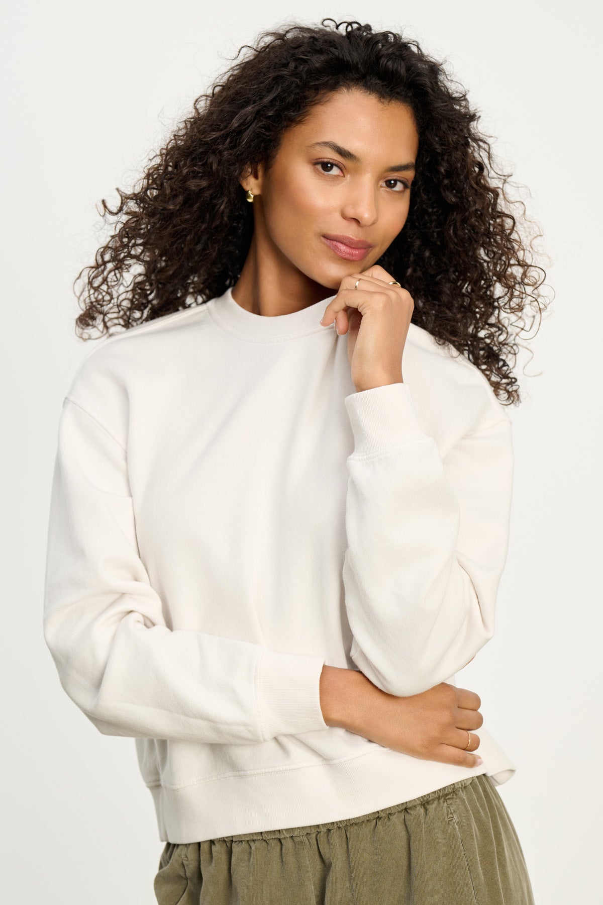 A woman with curly hair wears the YOKO SWEATSHIRT by Velvet by Graham & Spencer in off-white cotton fleece, paired with olive green pants, standing against a plain light background.-40431120711873