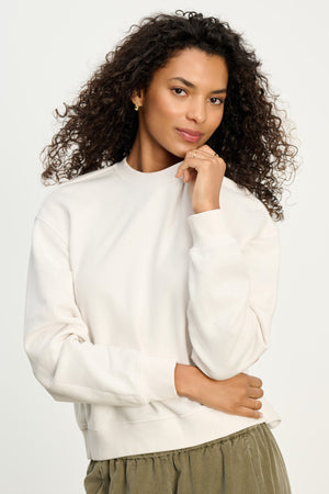 A woman with curly hair wears the YOKO SWEATSHIRT by Velvet by Graham & Spencer in off-white cotton fleece, paired with olive green pants, standing against a plain light background.