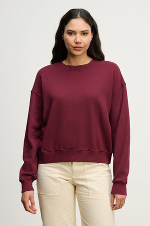 A woman faces forward against a plain light background, wearing Velvet by Graham & Spencer’s maroon YOKO SWEATSHIRT in cotton fleece paired with cream-colored pants.