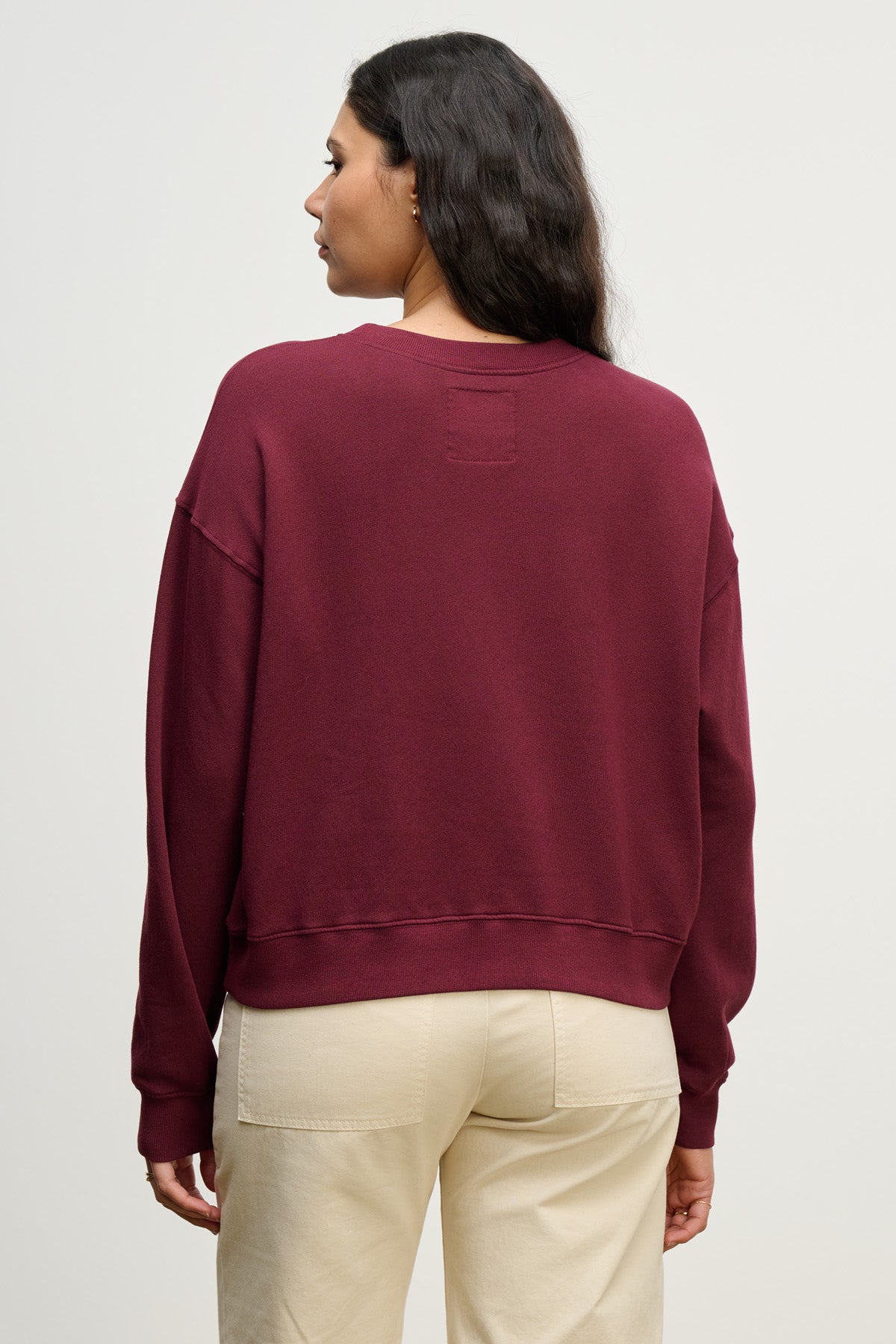 A woman with long dark hair is seen from the back, wearing the Velvet by Graham & Spencer YOKO SWEATSHIRT in maroon with a relaxed fit and beige pants, standing against a plain light background.