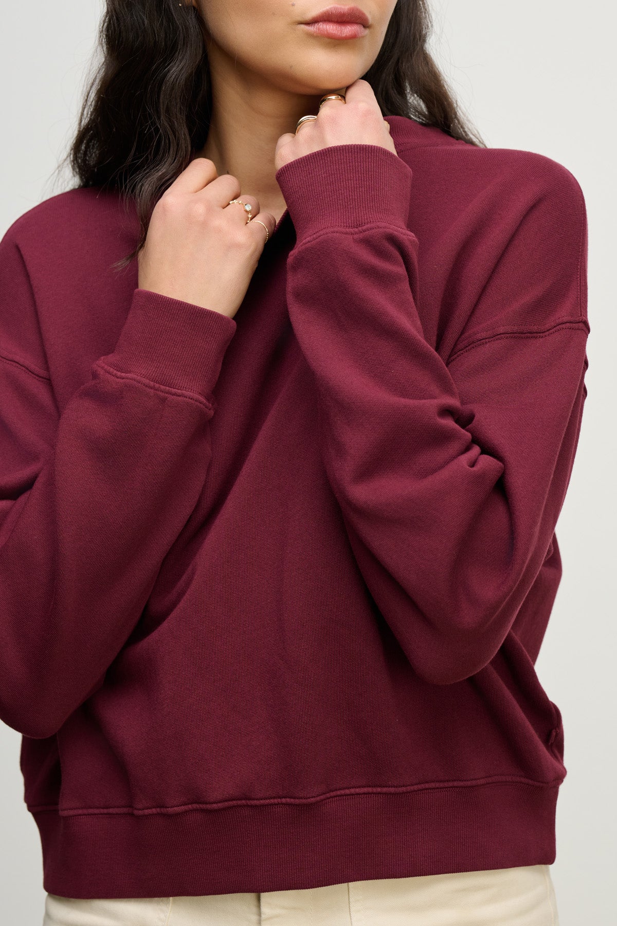 A person wears the Velvet by Graham & Spencer YOKO SWEATSHIRT in maroon, featuring long sleeves and a relaxed silhouette, paired with light-colored pants and holding the collar with both hands.