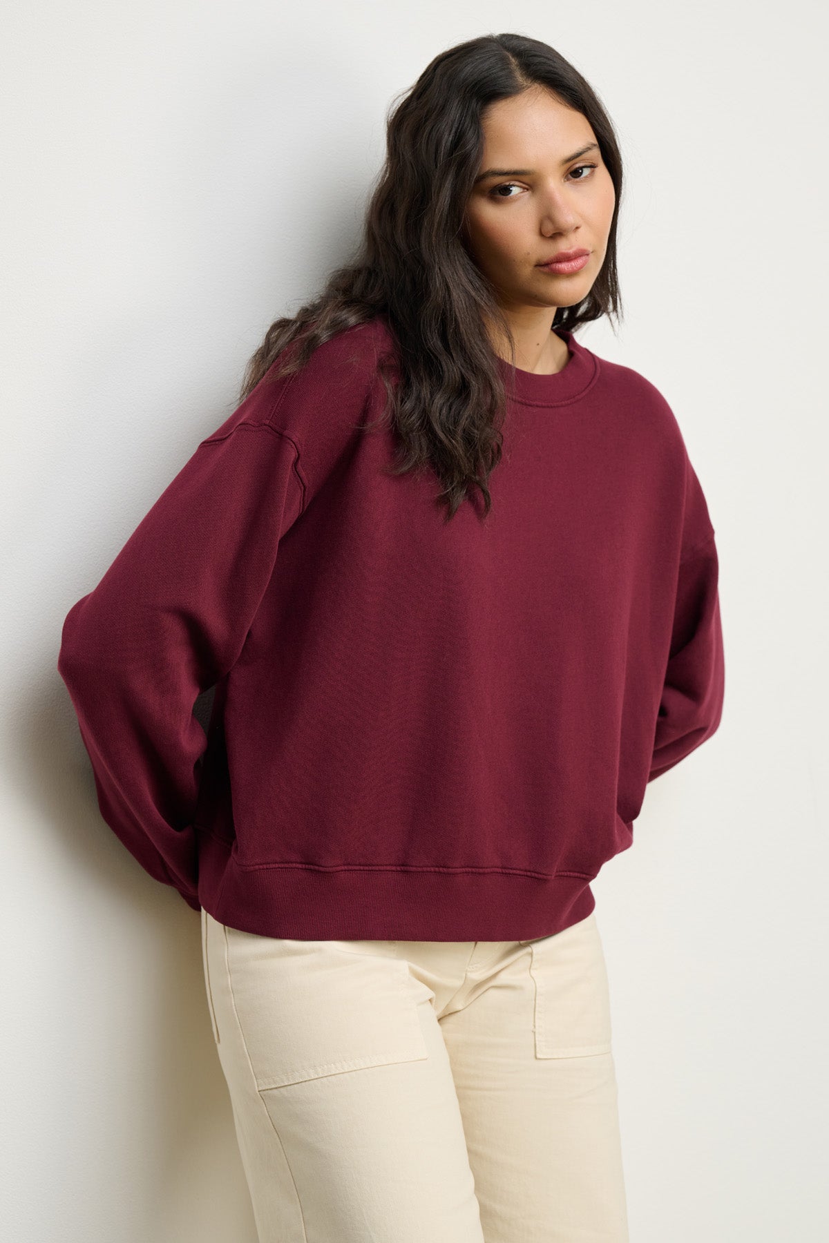 A person with long dark hair wears the Velvet by Graham & Spencer YOKO SWEATSHIRT in maroon cotton fleece with cream pants, standing against a plain white wall and looking slightly to the side with a relaxed silhouette.-40328370028737