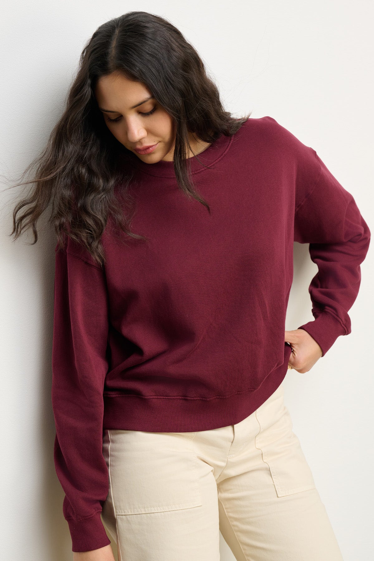 A woman with long dark hair wears the Velvet by Graham & Spencer YOKO SWEATSHIRT in maroon, made of soft cotton fleece, paired with light beige pants as she stands against a plain white wall, looking down.
