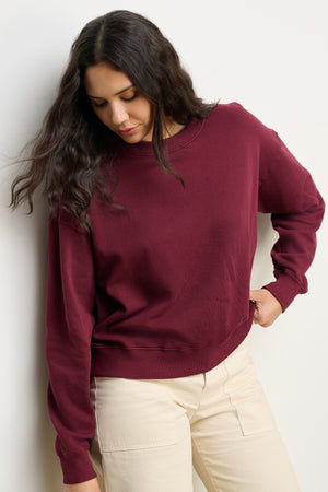 A woman with long dark hair wears the Velvet by Graham & Spencer YOKO SWEATSHIRT in maroon, made of soft cotton fleece, paired with light beige pants as she stands against a plain white wall, looking down.