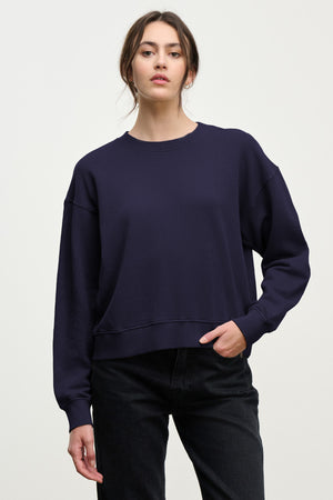 A person in a loose-fitting, long-sleeve YOKO SWEATSHIRT by Velvet by Graham & Spencer and black jeans poses against a plain light background, highlighting a relaxed silhouette.