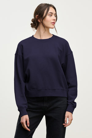 A woman in a loose-fitting YOKO SWEATSHIRT by Velvet by Graham & Spencer, made from navy blue cotton fleece, paired with black jeans, stands against a plain light background, looking to the side and highlighting the relaxed silhouette.