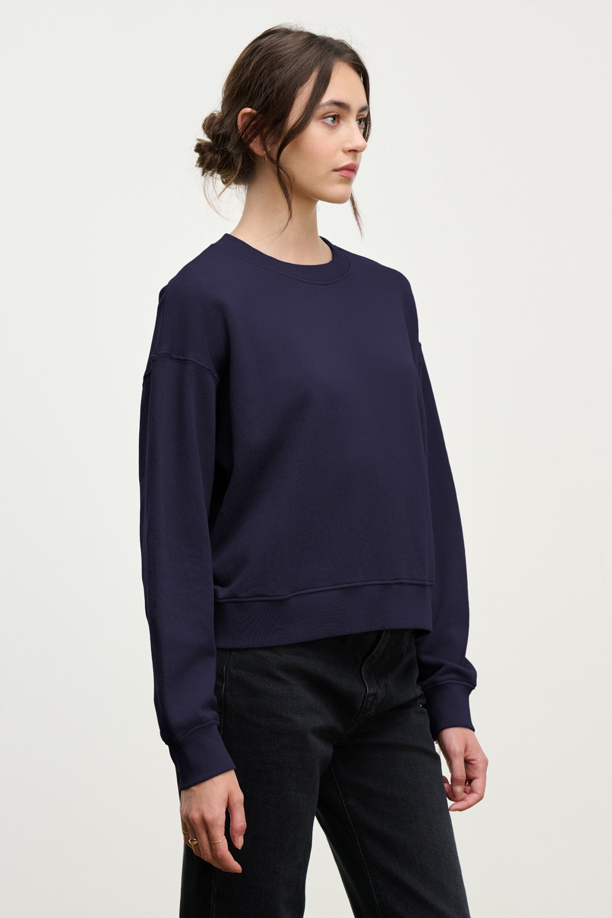 A woman with dark hair in a low bun wears the Velvet by Graham & Spencer YOKO SWEATSHIRT in navy cotton fleece and black jeans, highlighting its relaxed fit against a plain light background.