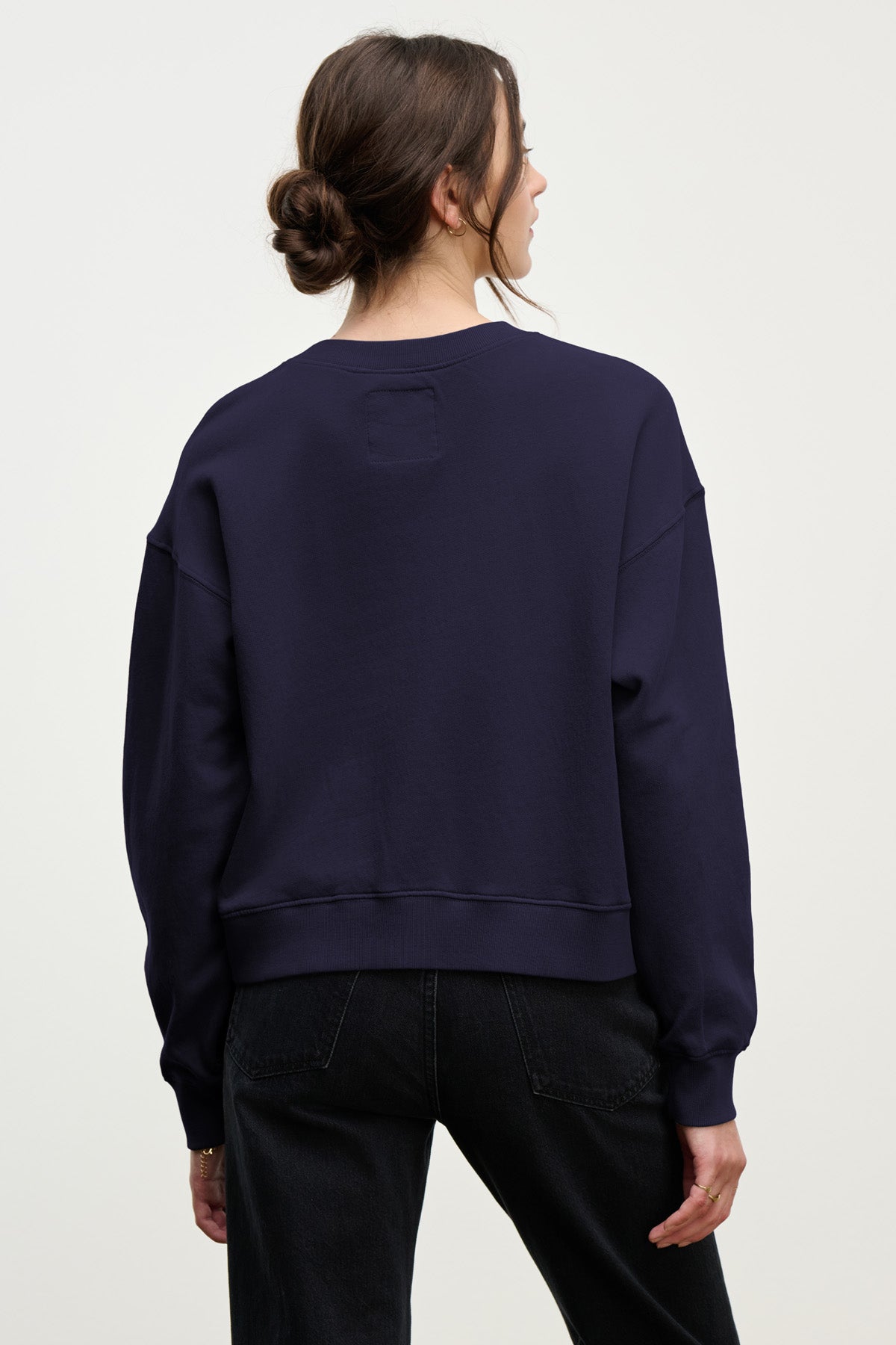 Woman with brown hair in a low bun wears the Velvet by Graham & Spencer YOKO SWEATSHIRT in dark blue cotton fleece and black jeans, standing with her back to the camera against a plain light background.-40431164096705