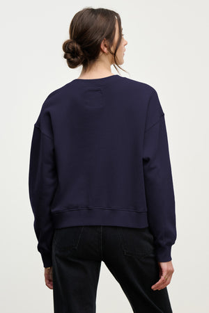 Woman with brown hair in a low bun wears the Velvet by Graham & Spencer YOKO SWEATSHIRT in dark blue cotton fleece and black jeans, standing with her back to the camera against a plain light background.