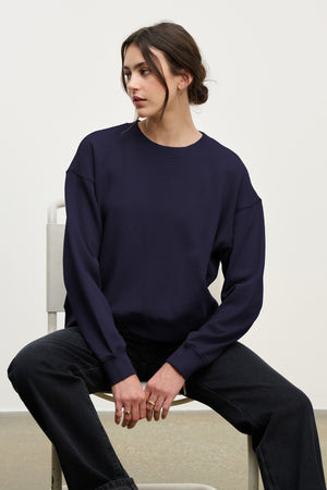 A person with dark hair in a low bun sits on a white chair, wearing the Velvet by Graham & Spencer YOKO SWEATSHIRT in a relaxed fit and black jeans, gazing to the side against a simple background.