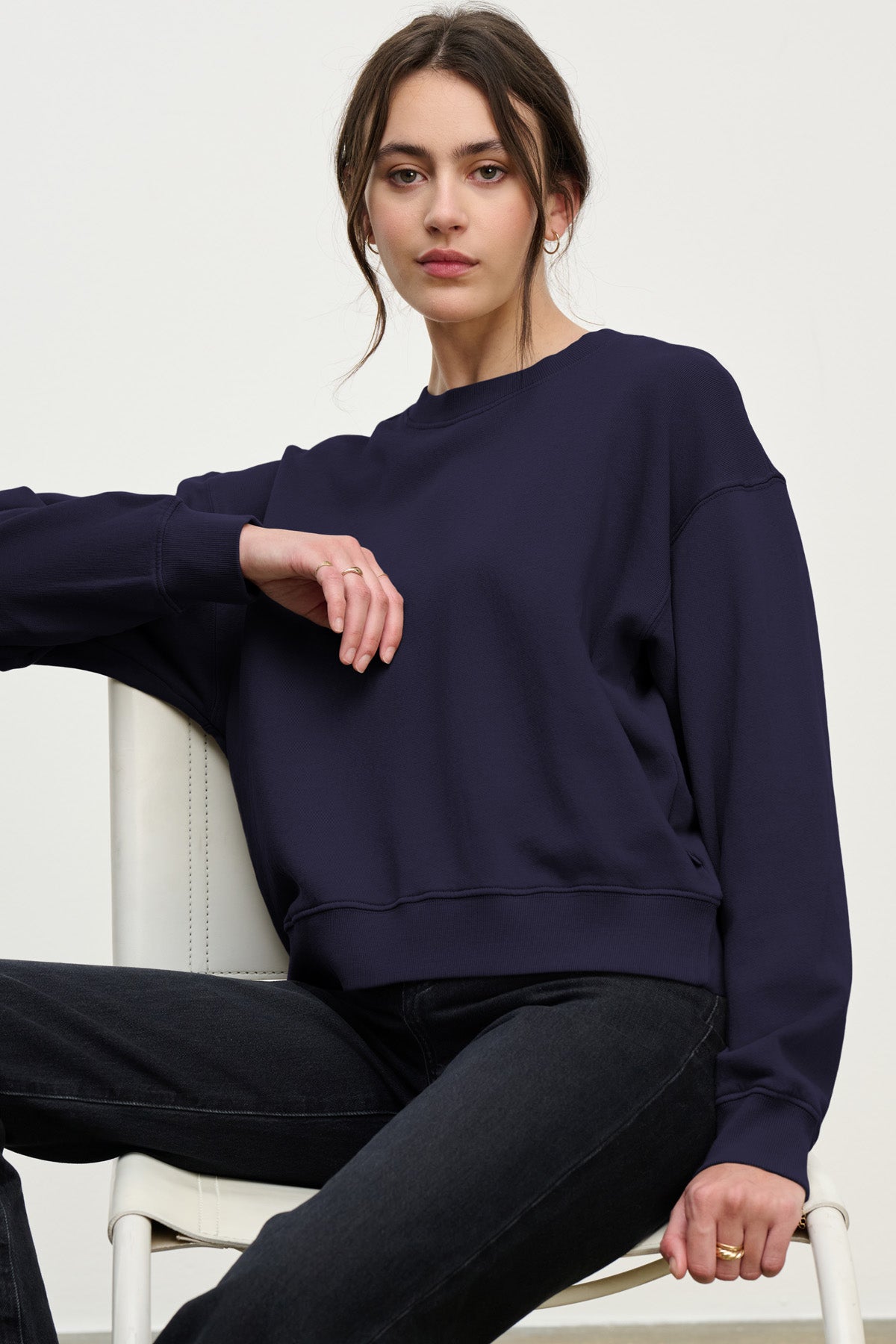 A woman sits on a white chair in the Velvet by Graham & Spencer YOKO SWEATSHIRT—navy blue cotton fleece—paired with black jeans, her relaxed silhouette and calm, neutral expression completing the look.-40431163998401