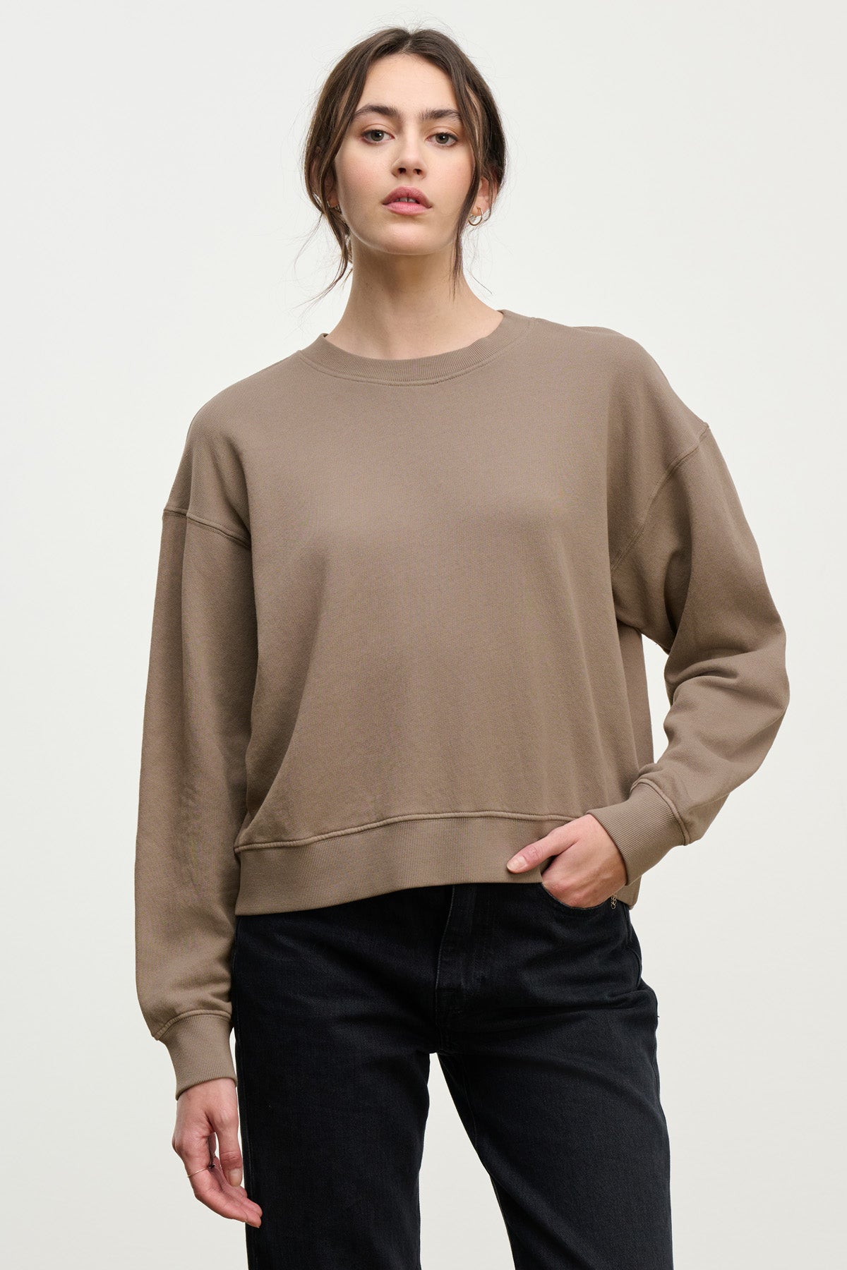 A woman wears a loose taupe YOKO SWEATSHIRT by Velvet by Graham & Spencer in soft cotton fleece with black jeans, standing against a plain light background and looking at the camera.-40328390770881