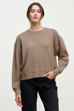 A woman wears a loose taupe YOKO SWEATSHIRT by Velvet by Graham & Spencer in soft cotton fleece with black jeans, standing against a plain light background and looking at the camera.